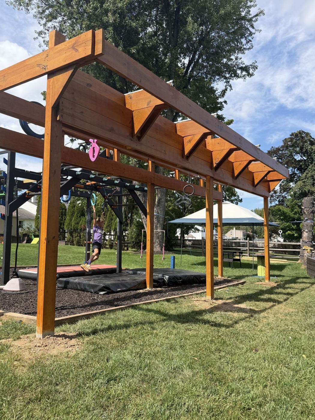 Finished stained-wood backyard ninja course with steel bars, rings and hanging grips over a rubber-mulch landing bed, a young ninja swinging across