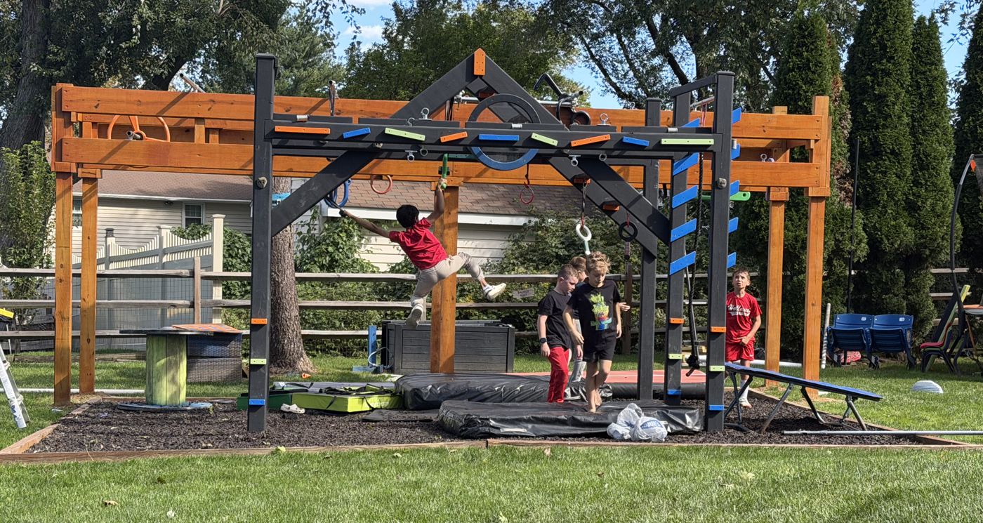 Kids training together on a backyard ninja course, a stained wooden rig behind a black steel frame